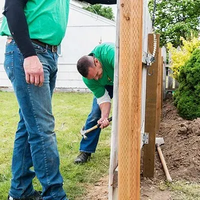 Two men installing a wooden fence in a backyard. One man hammers while the other looks on.