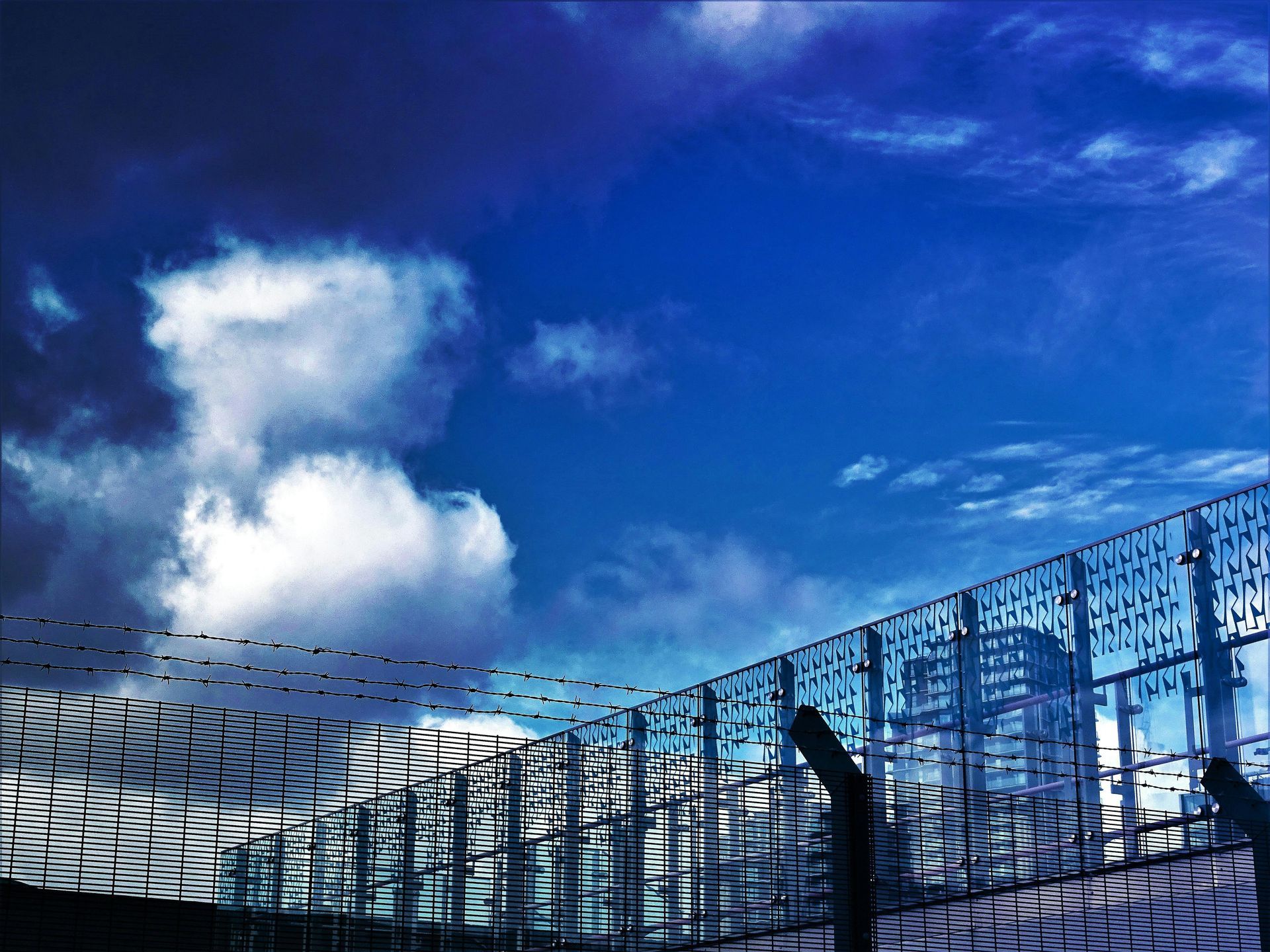 Blue sky with white clouds above a tall, chain-link fence with barbed wire. City buildings are visible behind the fence.