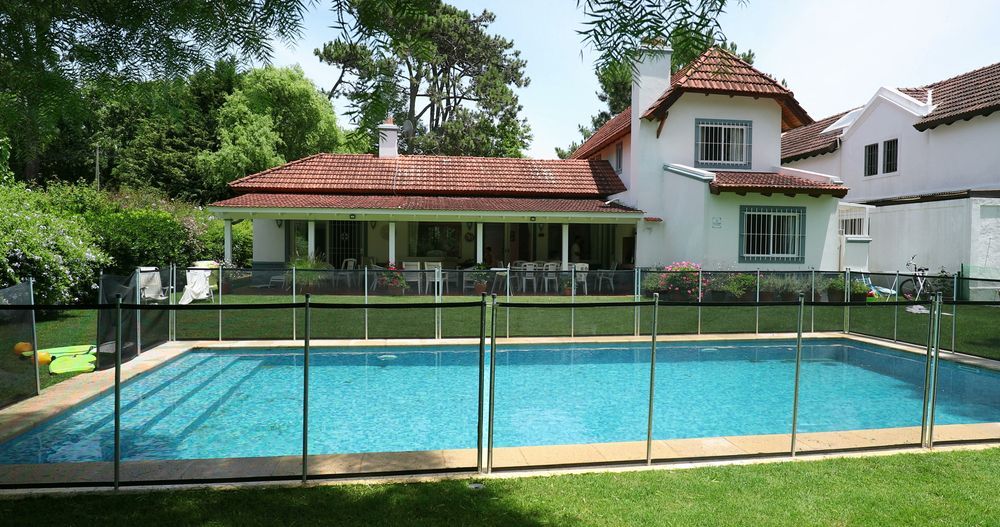 A white house with a red-tiled roof overlooks a rectangular pool surrounded by a black mesh fence. Lush green trees border the scene.