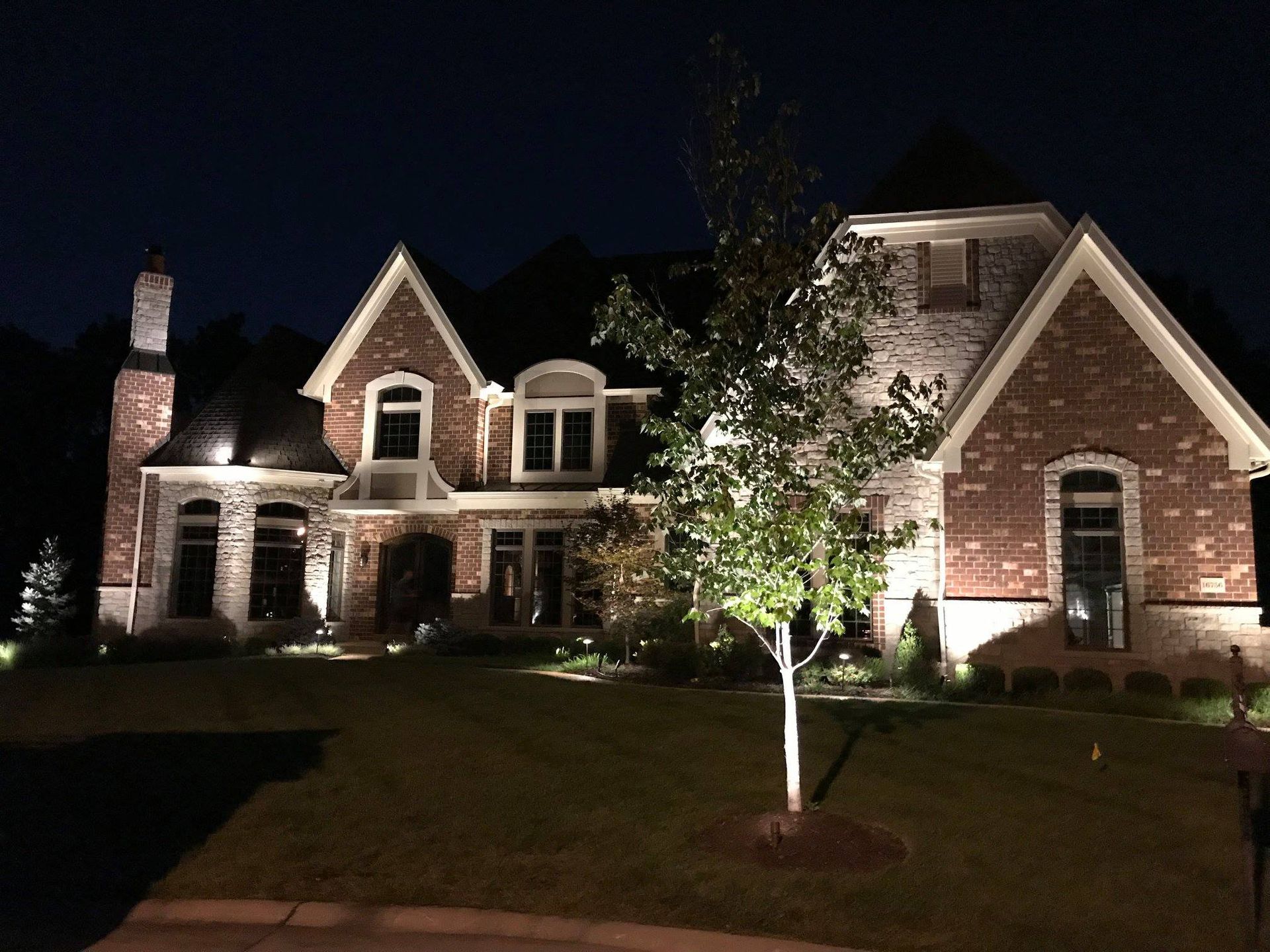Large brick-and-stone house at night with exterior lights illuminating the front yard and trees