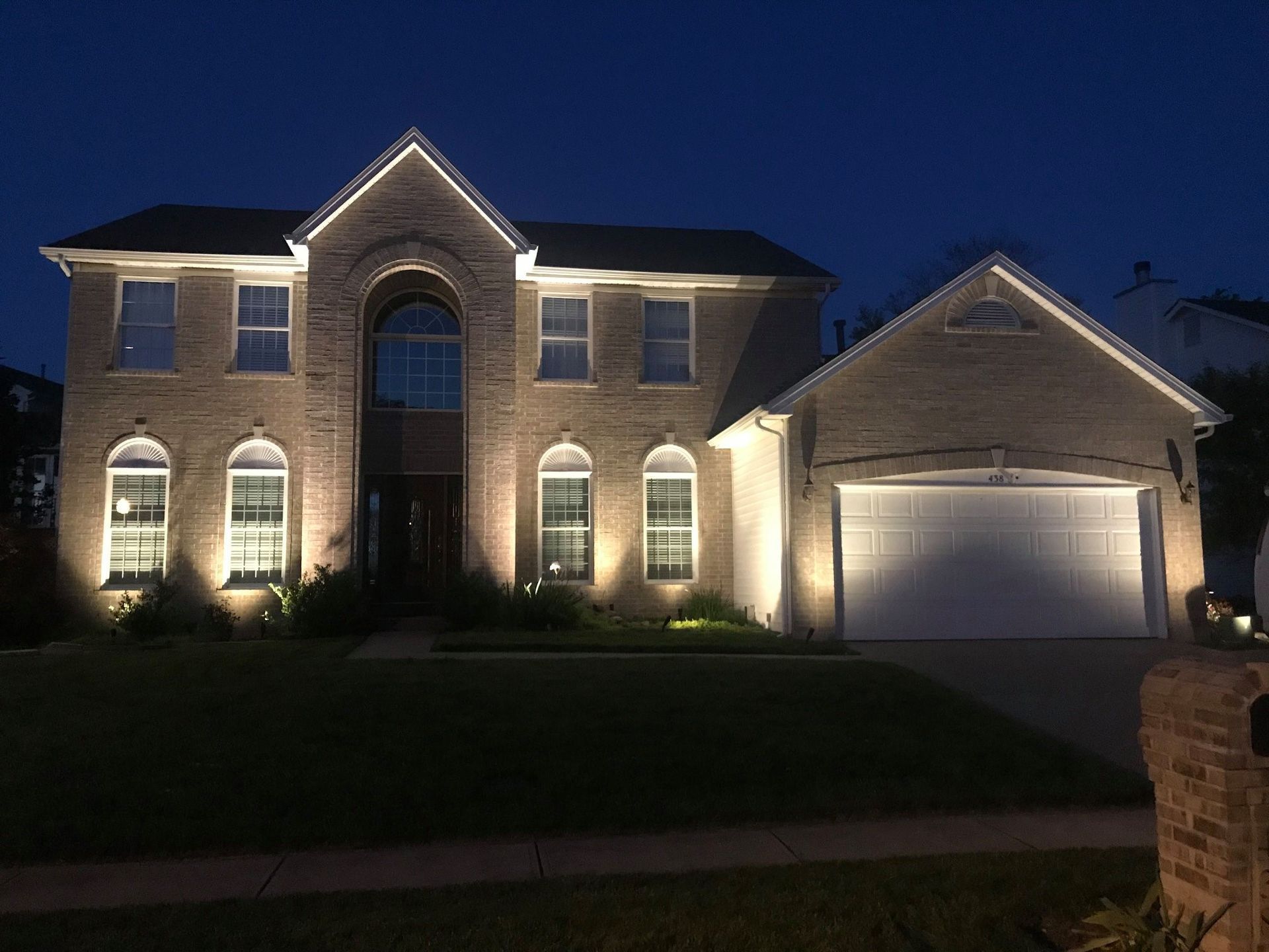 Two-story brick house at dusk, lit from below, with a garage and manicured front lawn