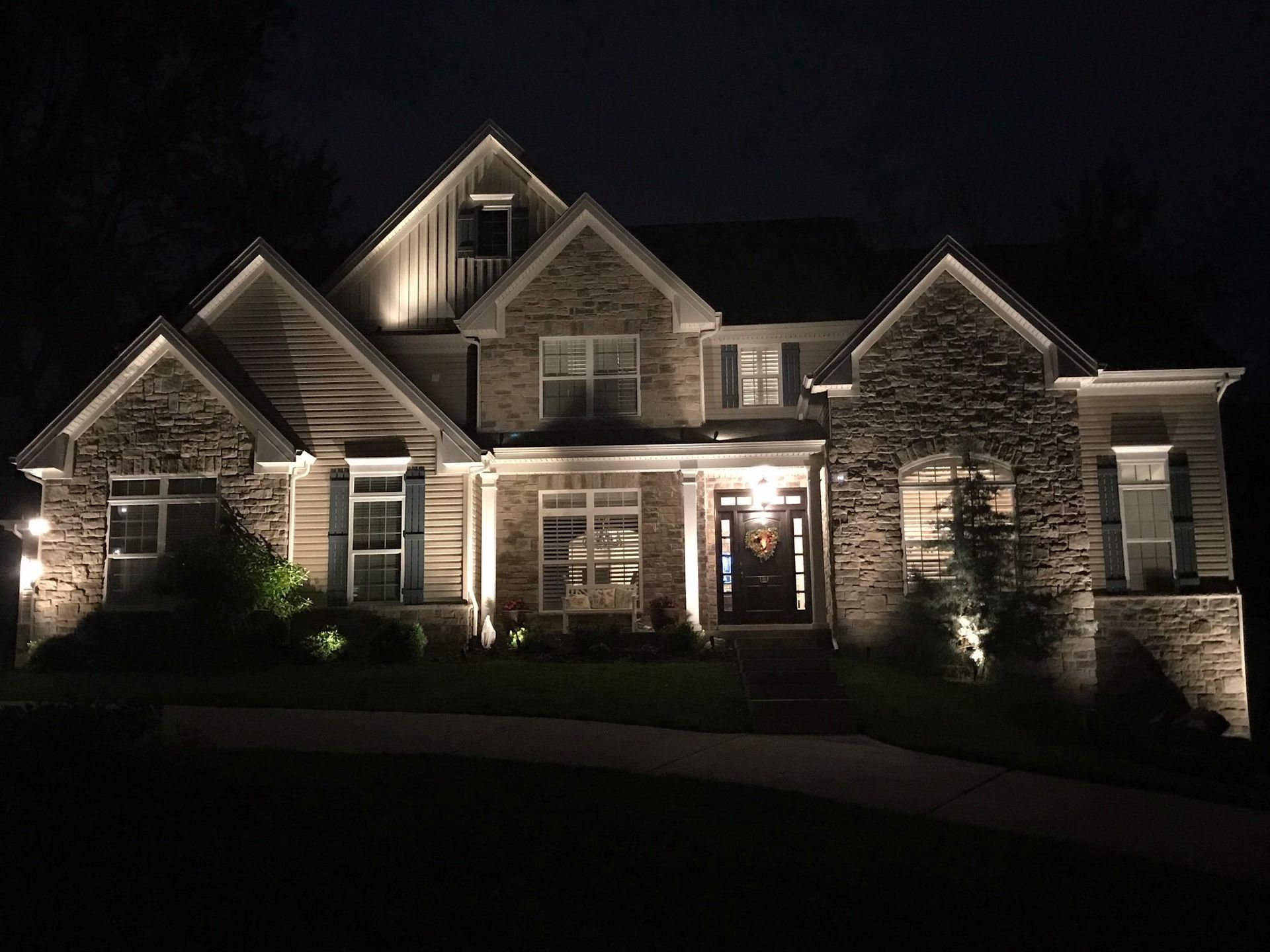 Nighttime view of a large stone-and-siding house lit by warm exterior lights