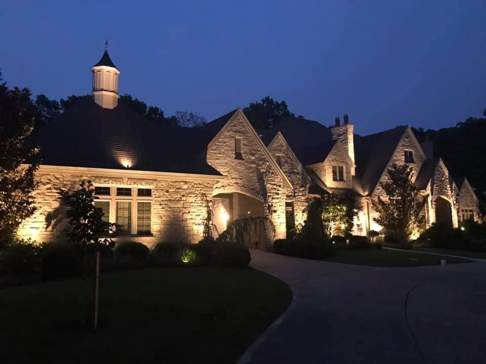 Large stone house with lit windows, purple-lit trees, and a landscaped garden at dusk