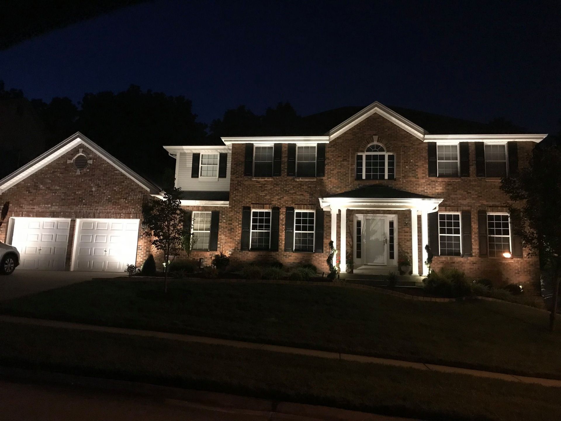 Brick house and garage at night, brightly lit with white trim and front porch pillars.