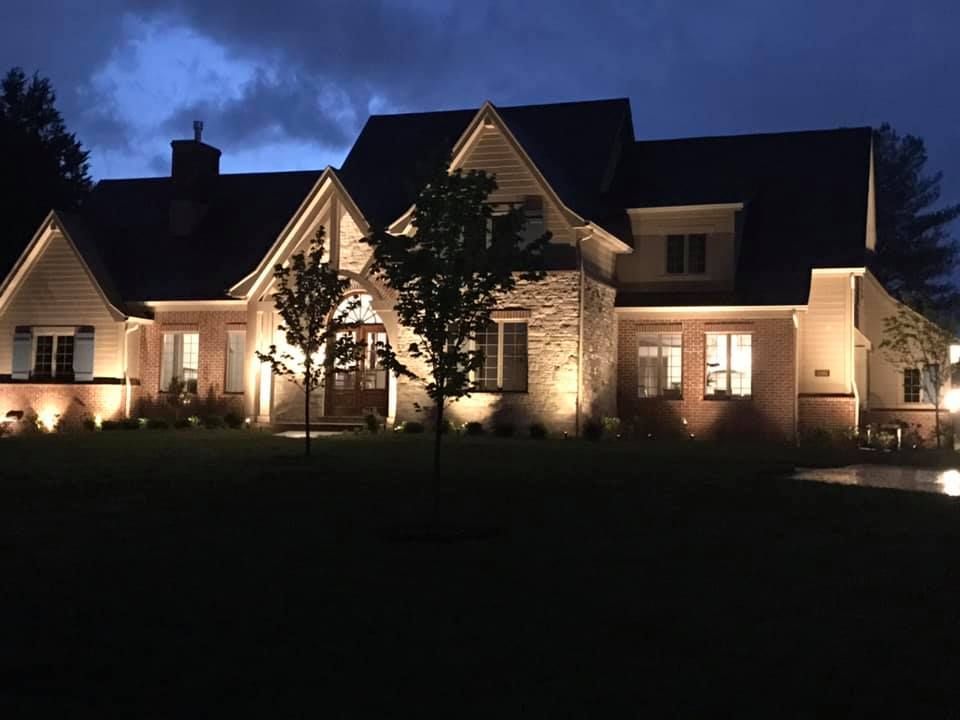 Large suburban house at night, warmly lit windows and front yard against a dark sky.