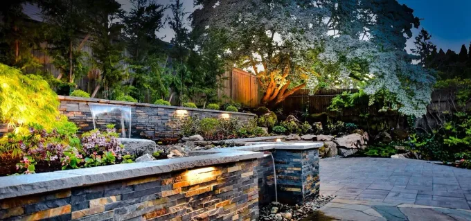 Illuminated stone patio and garden terrace with steps, shrubs, and trees at dusk