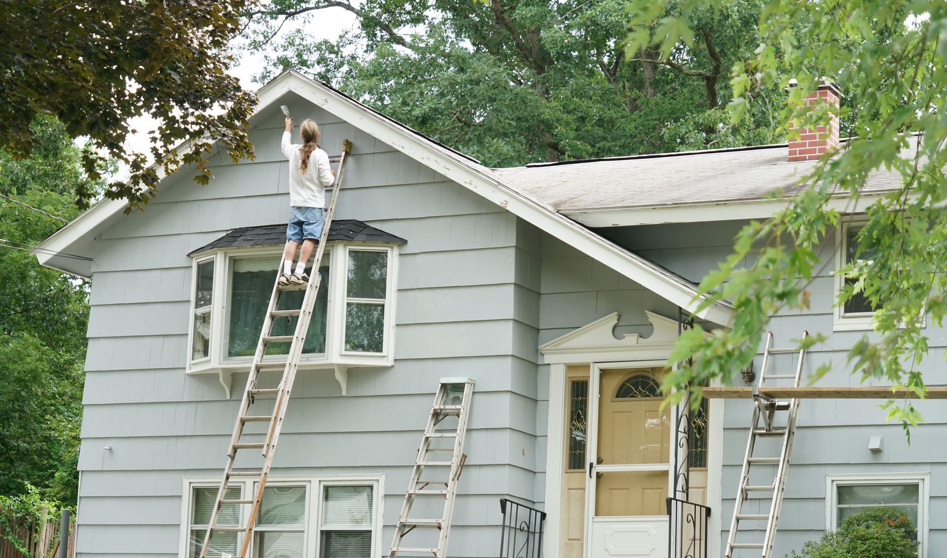 Person painting the trim of a light blue house from a tall ladder on a sunny day.