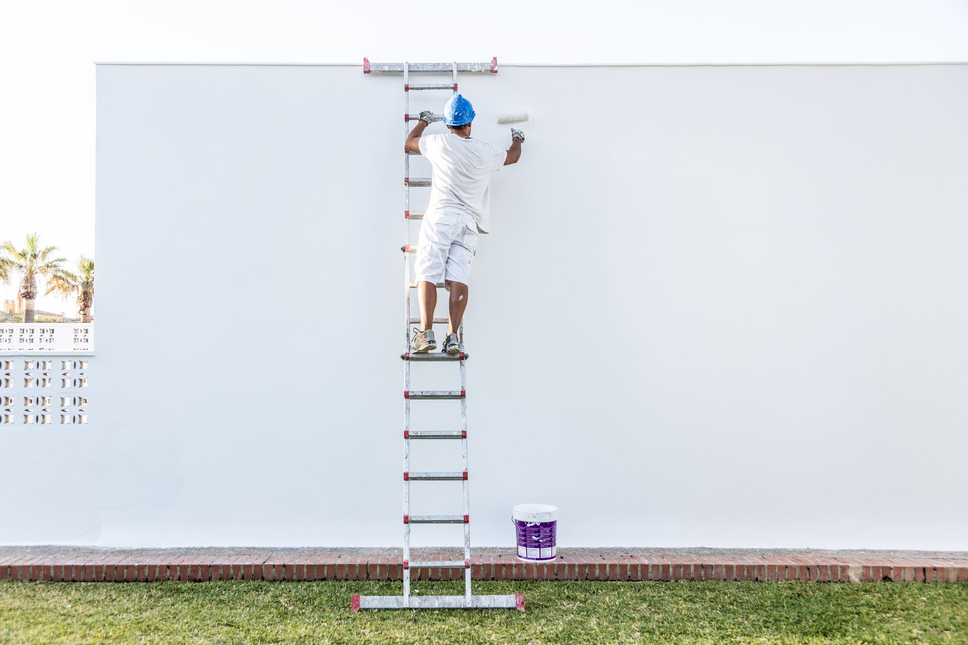 A painter is applying white paint to an exterior wall, with a paint bucket below.