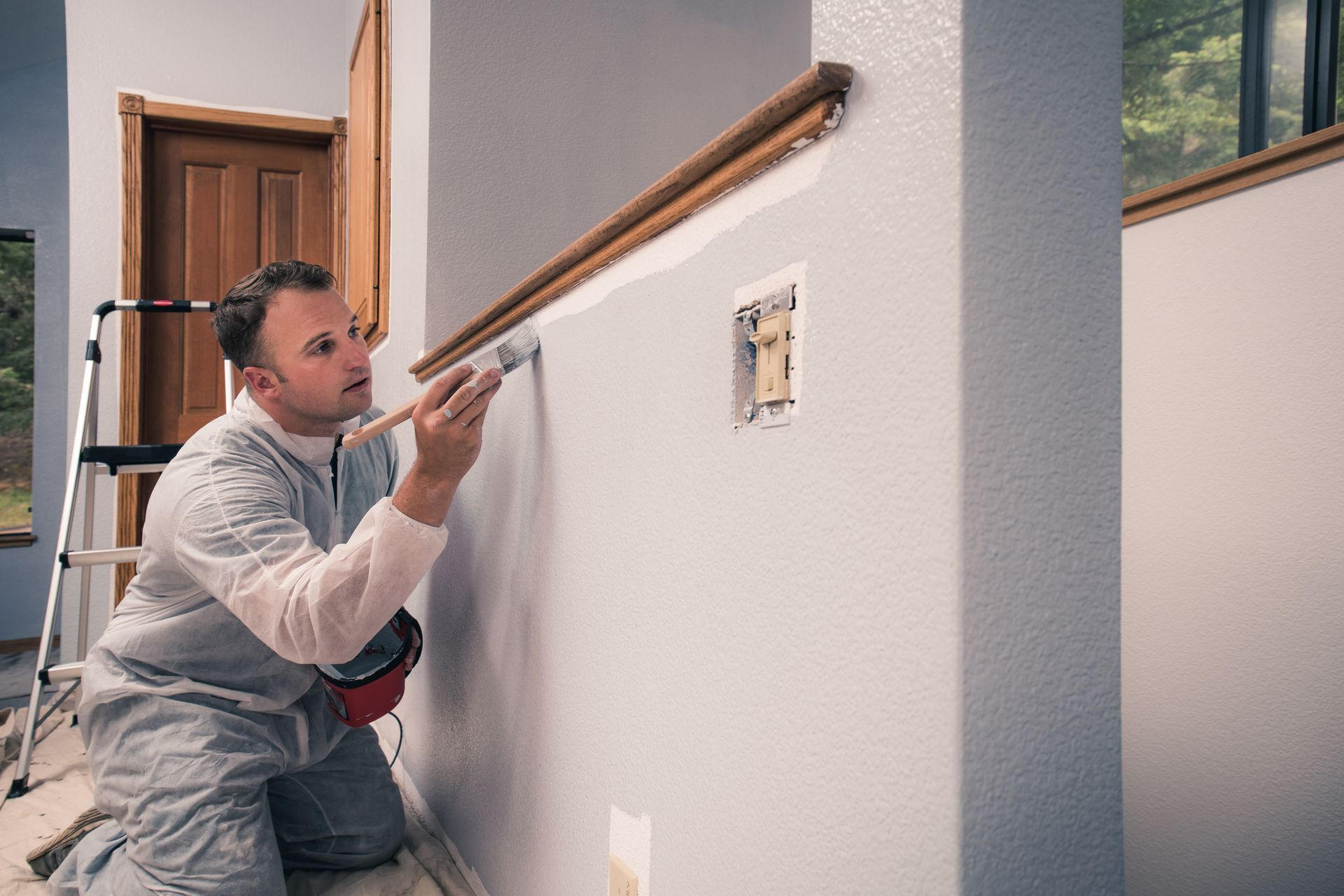 Man painting a wall with a brush, wearing a jumpsuit. Gray wall with wood trim in a room.
