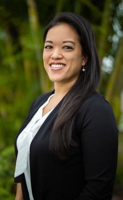 Woman with long dark hair smiles, wearing a black blazer over a white blouse, in front of greenery.