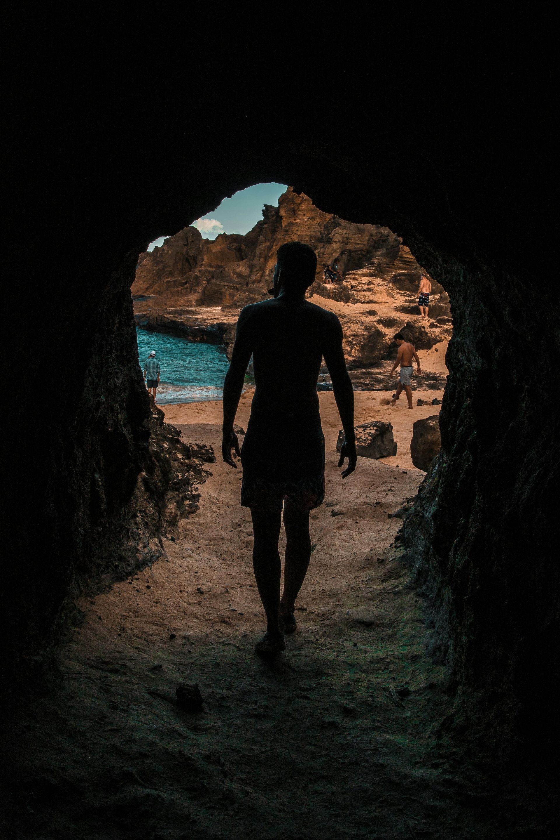 Silhouette of a person walking from a dark cave towards a sunny beach and ocean.
