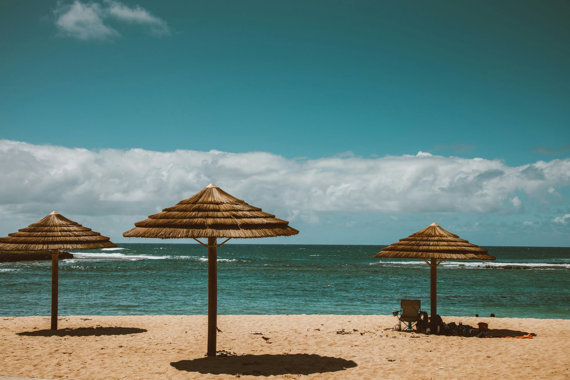 Three straw umbrellas on a sandy beach, turquoise water, blue sky with clouds.