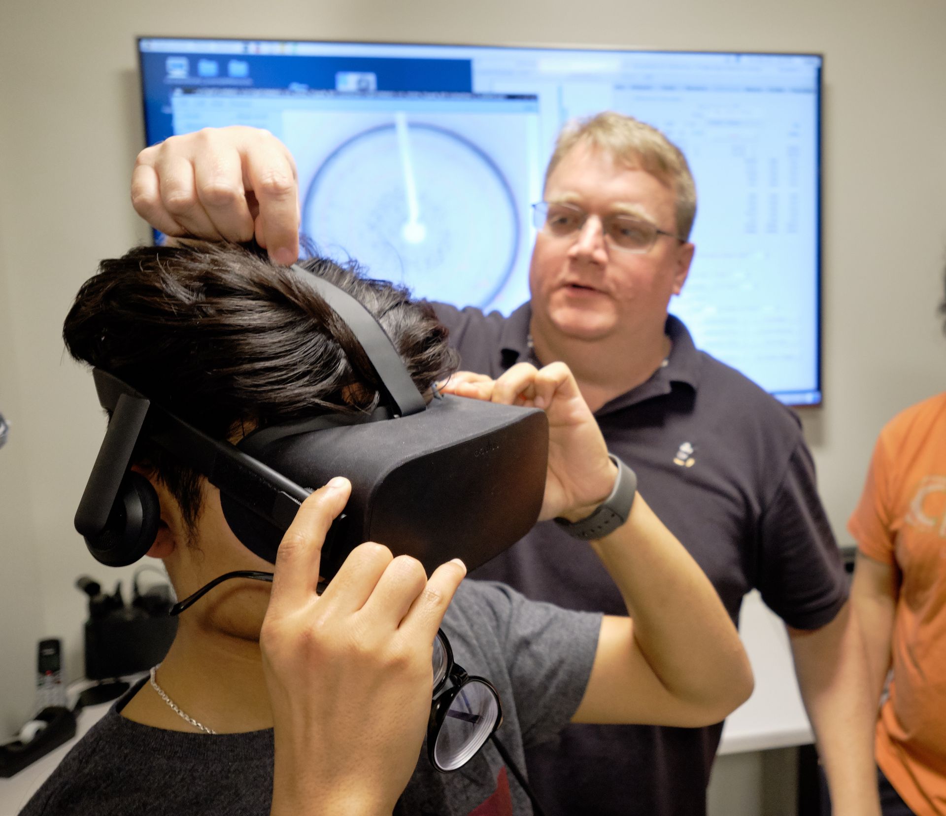 Man adjusting a VR headset on a person. White wall, computer screen in background.