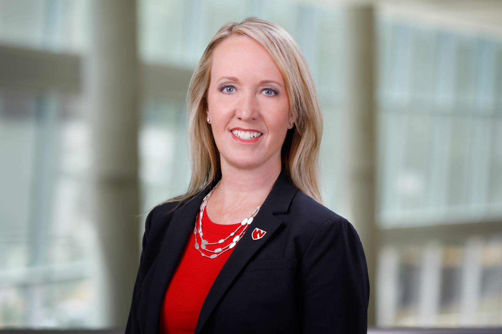 Woman with blonde hair wearing a red top and black blazer, smiling in an office setting.