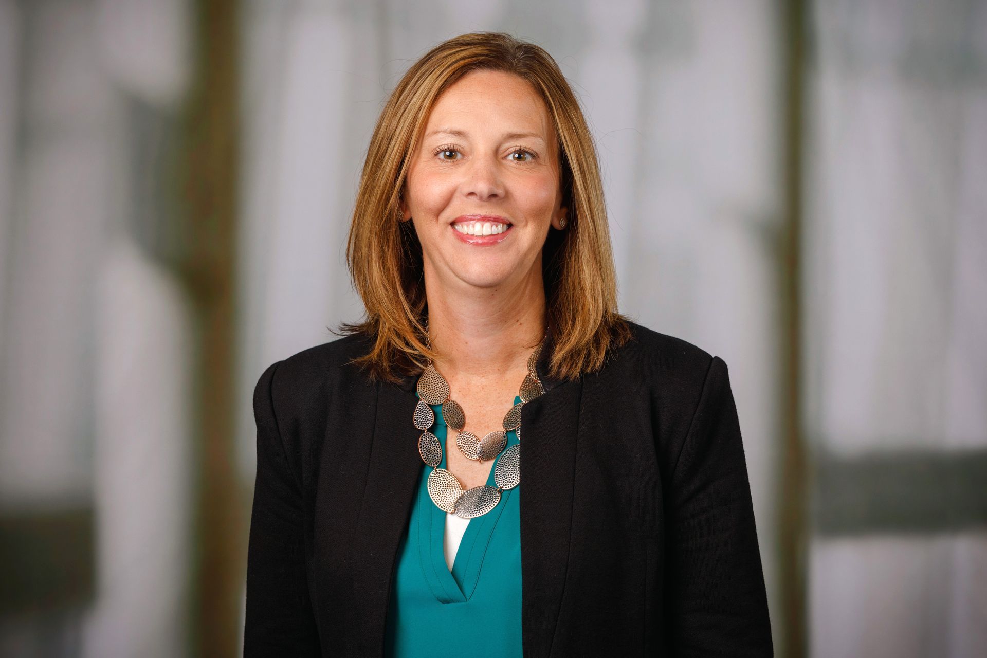 Woman with brown hair smiles, wearing a black blazer, teal top, and necklace. Blurred background.