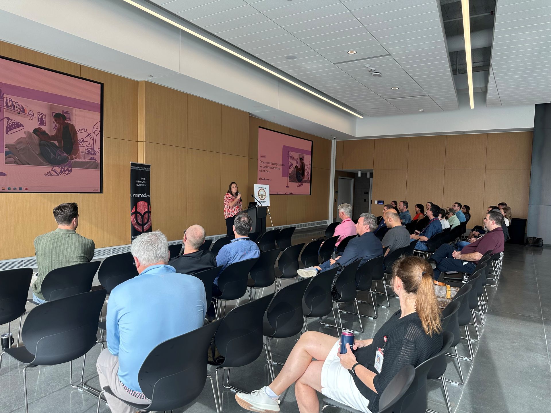 Audience in a conference room listening to a presenter, with art on the walls.