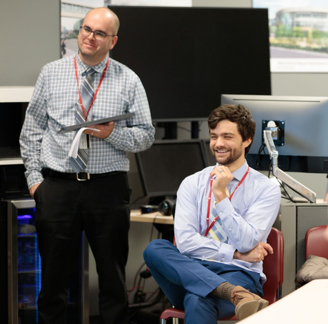 Two men in a tech setting, one standing with tablet, the other seated and smiling, wearing lanyards.