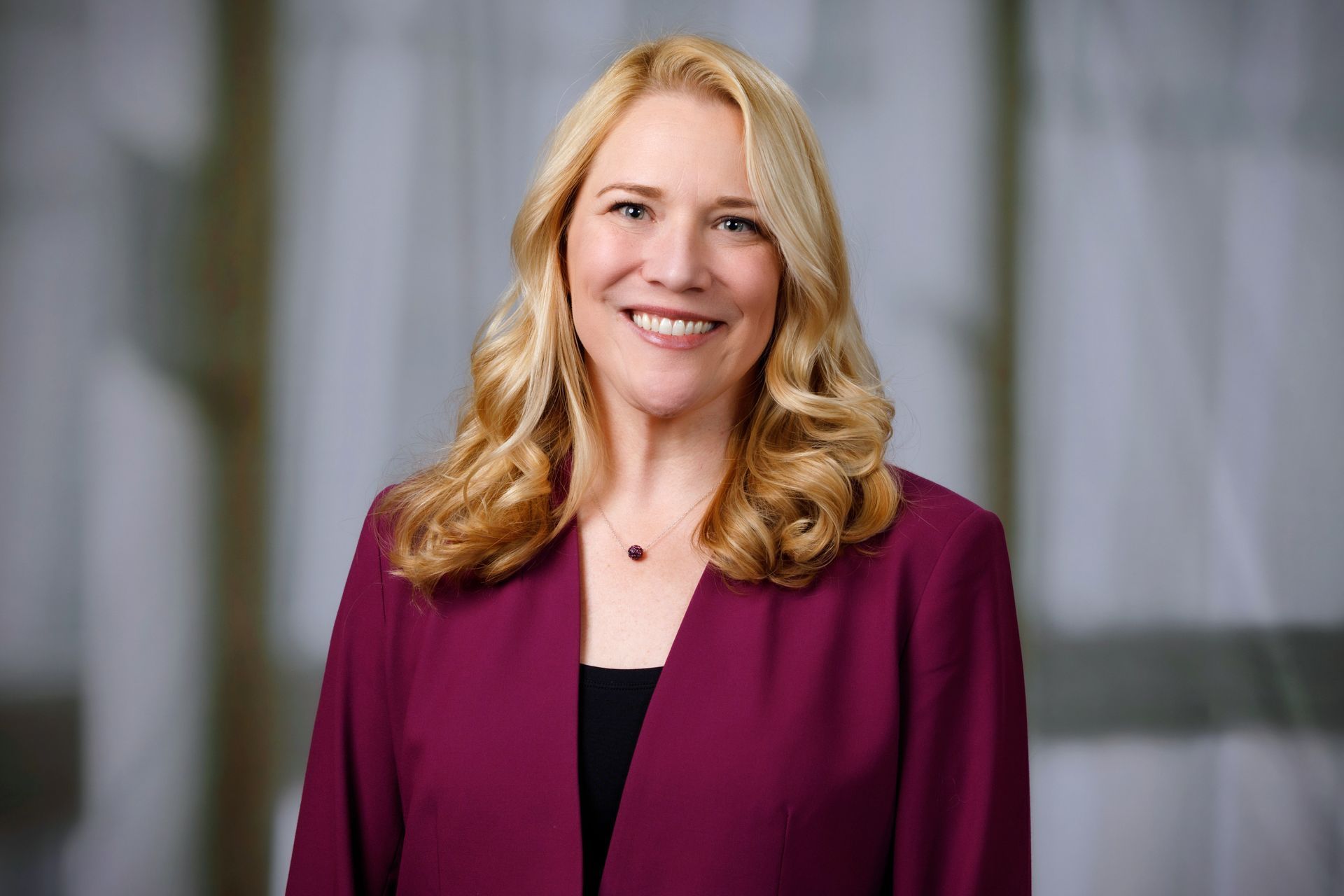 Woman with blonde hair wearing a maroon blazer smiles at the camera in front of a blurred background.