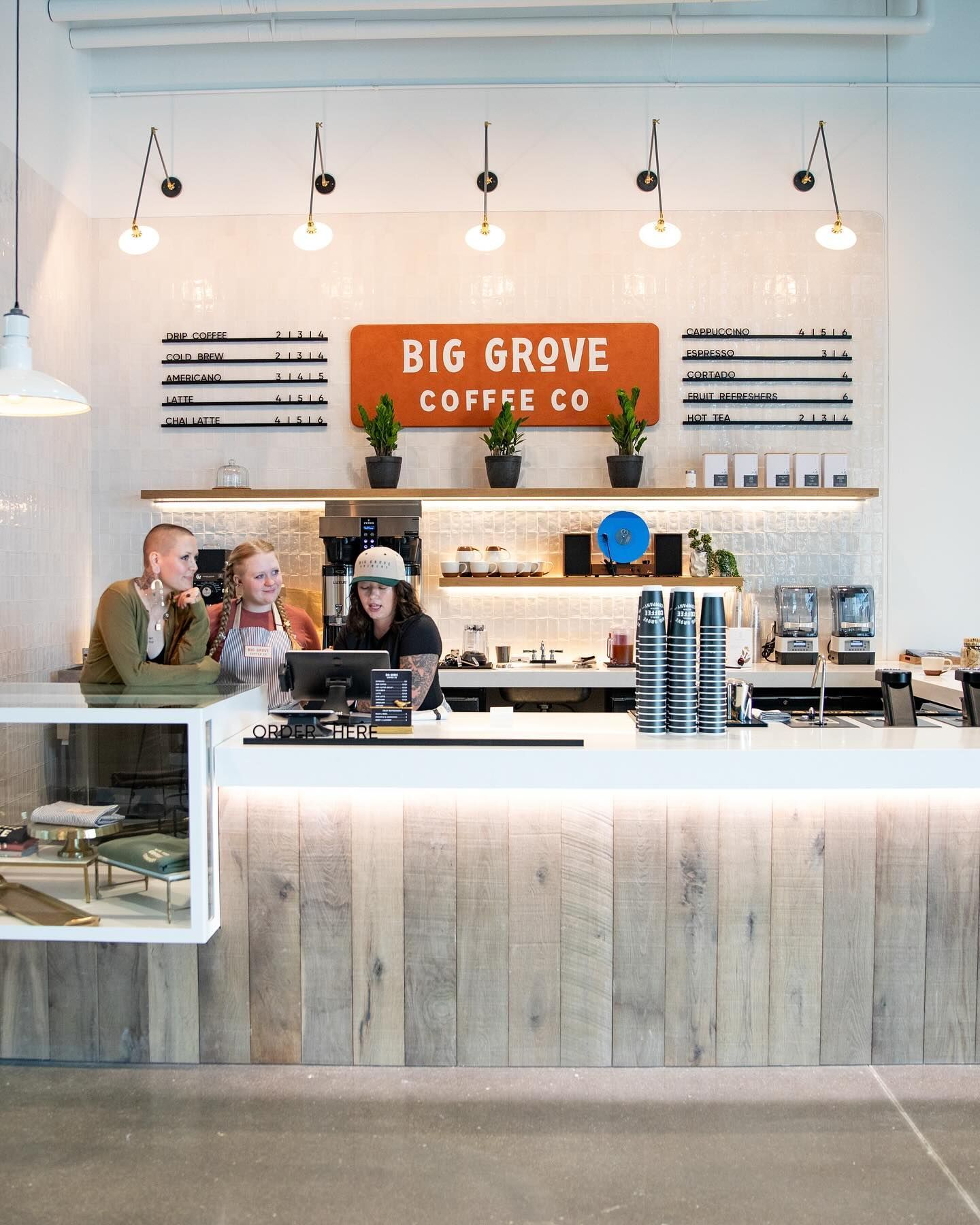 Coffee shop interior: barista serving customers at a counter with wooden paneling; orange Big Grove sign.