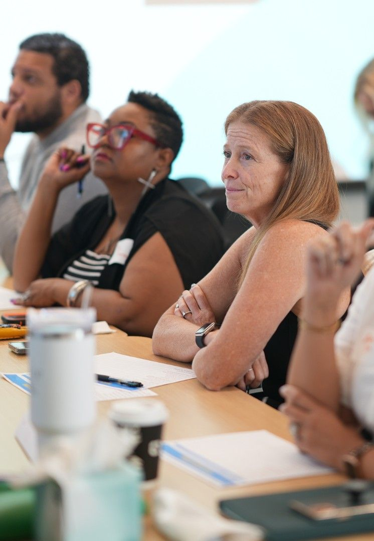 People seated at a table, attending a meeting. A woman in the foreground has reddish hair, listening intently.