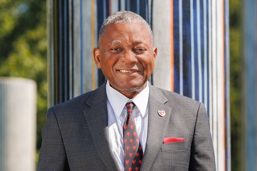 Smiling man in a suit, red tie, and pocket square, in front of a striped pillar outdoors.