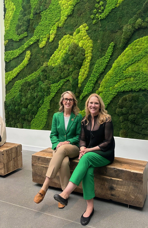 Two women sitting on a wooden bench in front of a green moss wall.