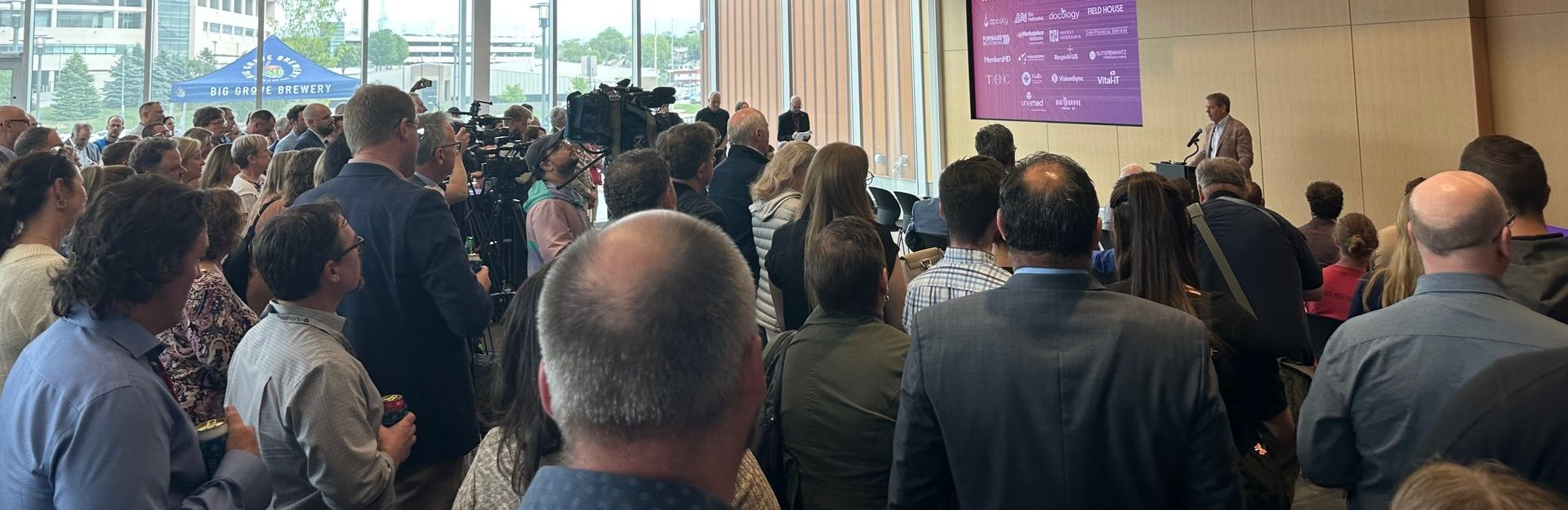 A packed conference room with a speaker at the front. Attendees listen and observe.