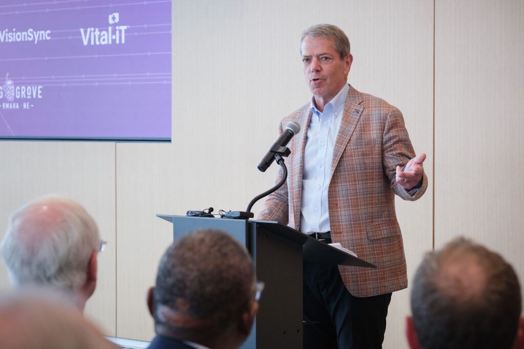 Man in blazer speaks at a podium, gesturing with his right hand, with an audience in front.