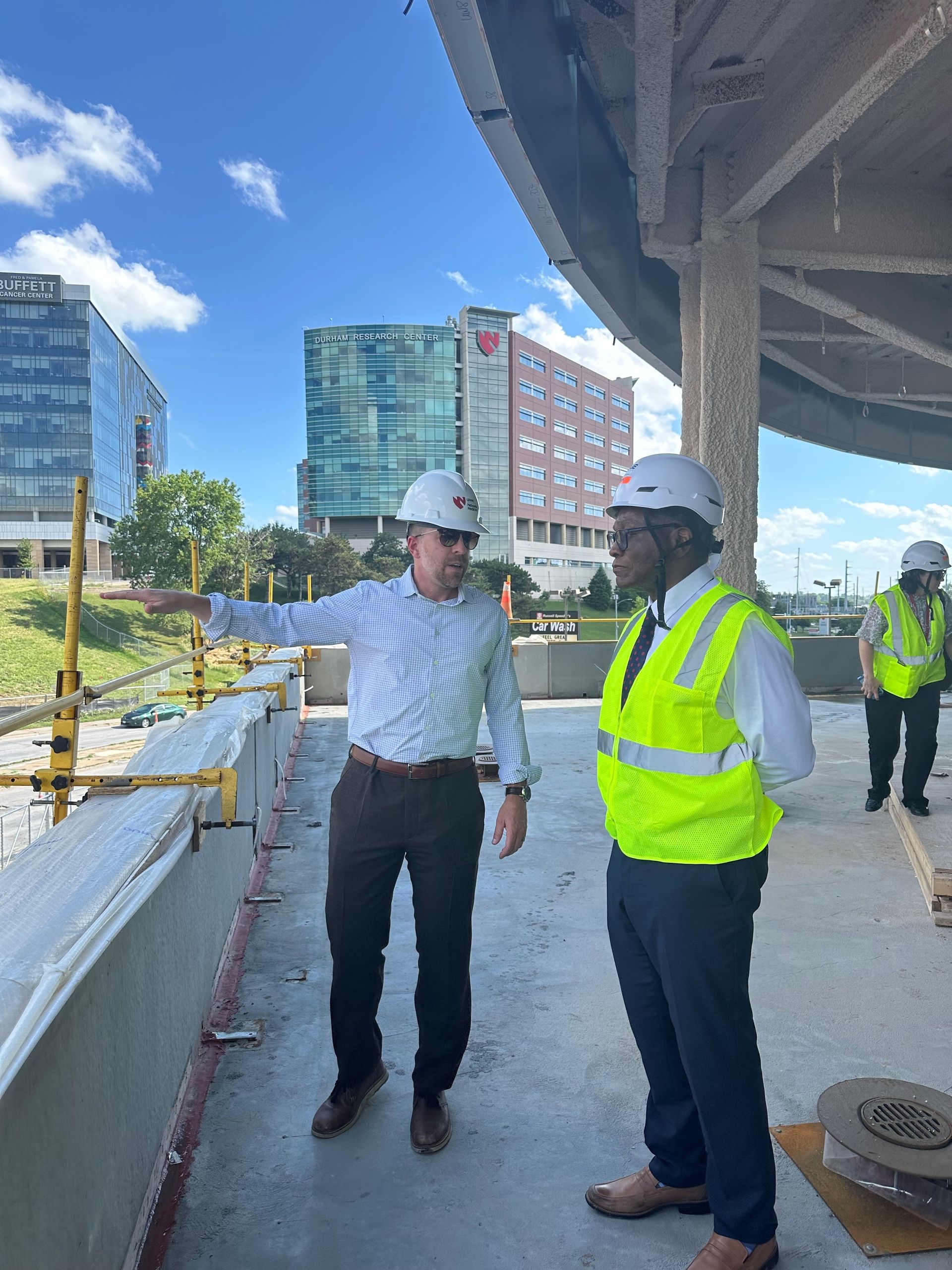 Two men in hard hats and vests on a construction site, one pointing to the distance.