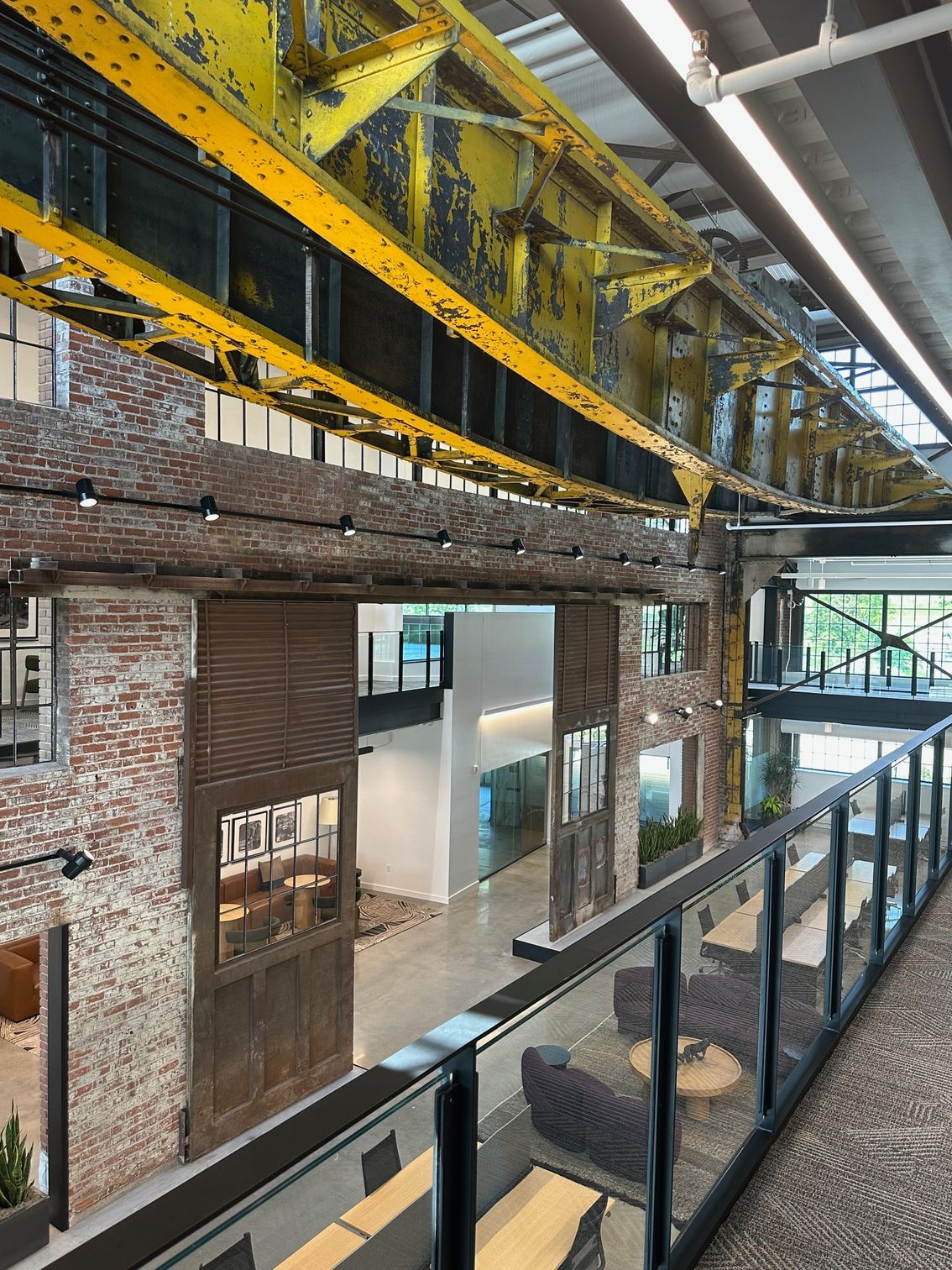 Interior view of a modern industrial building with exposed brick and yellow steel beams, overlooking a seating area.