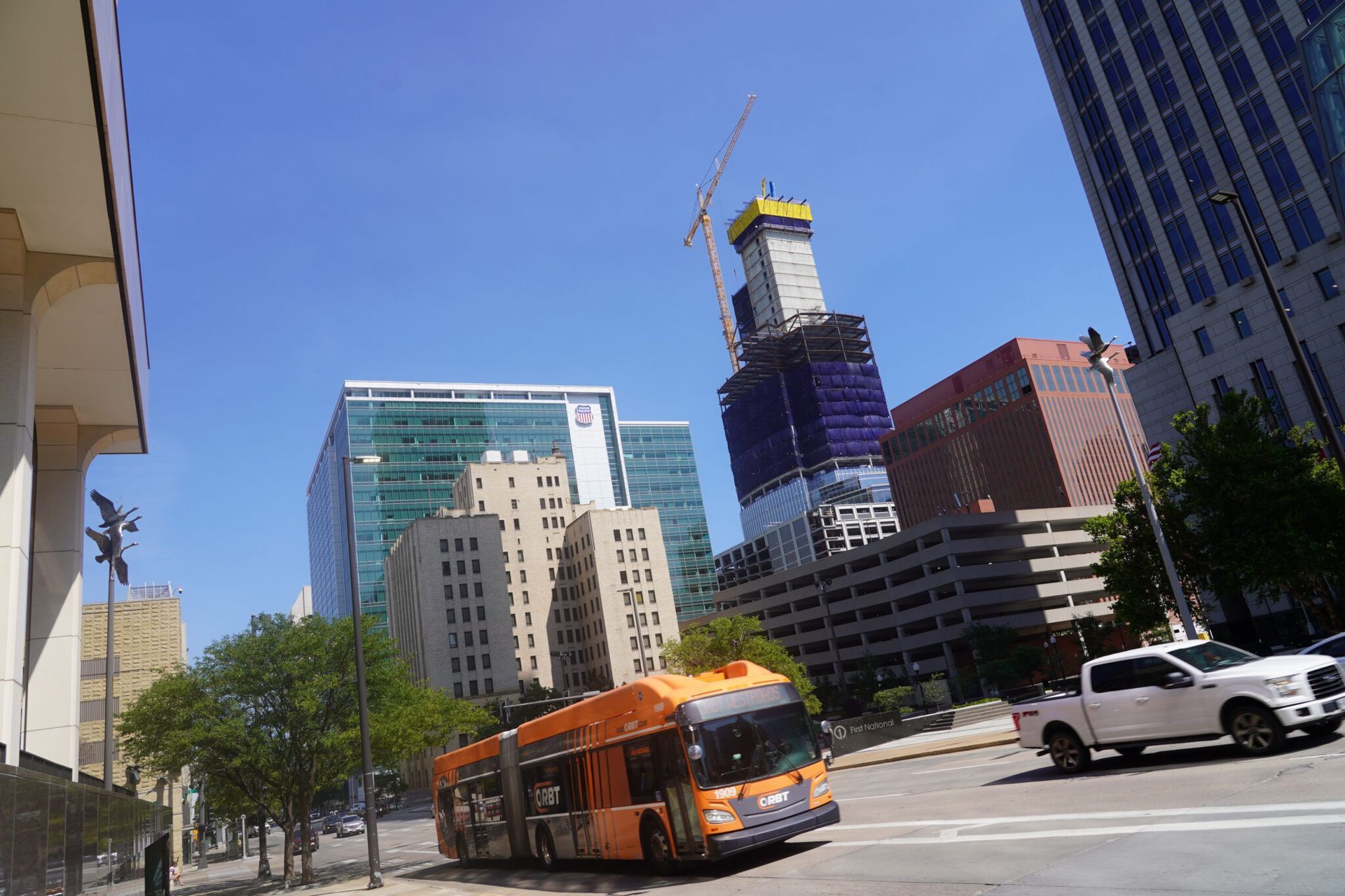 An orange bus drives on a city street with skyscrapers and construction crane in the background on a sunny day.