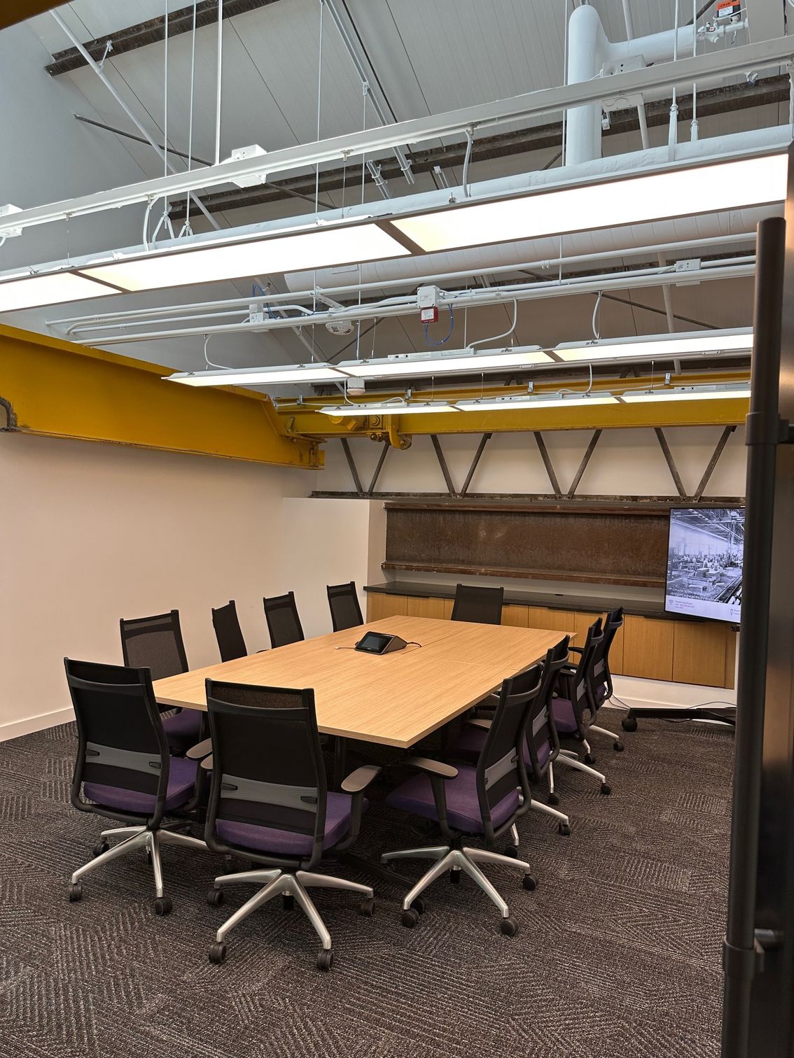 Conference room with a rectangular table, black chairs with purple accents, and a mounted screen.