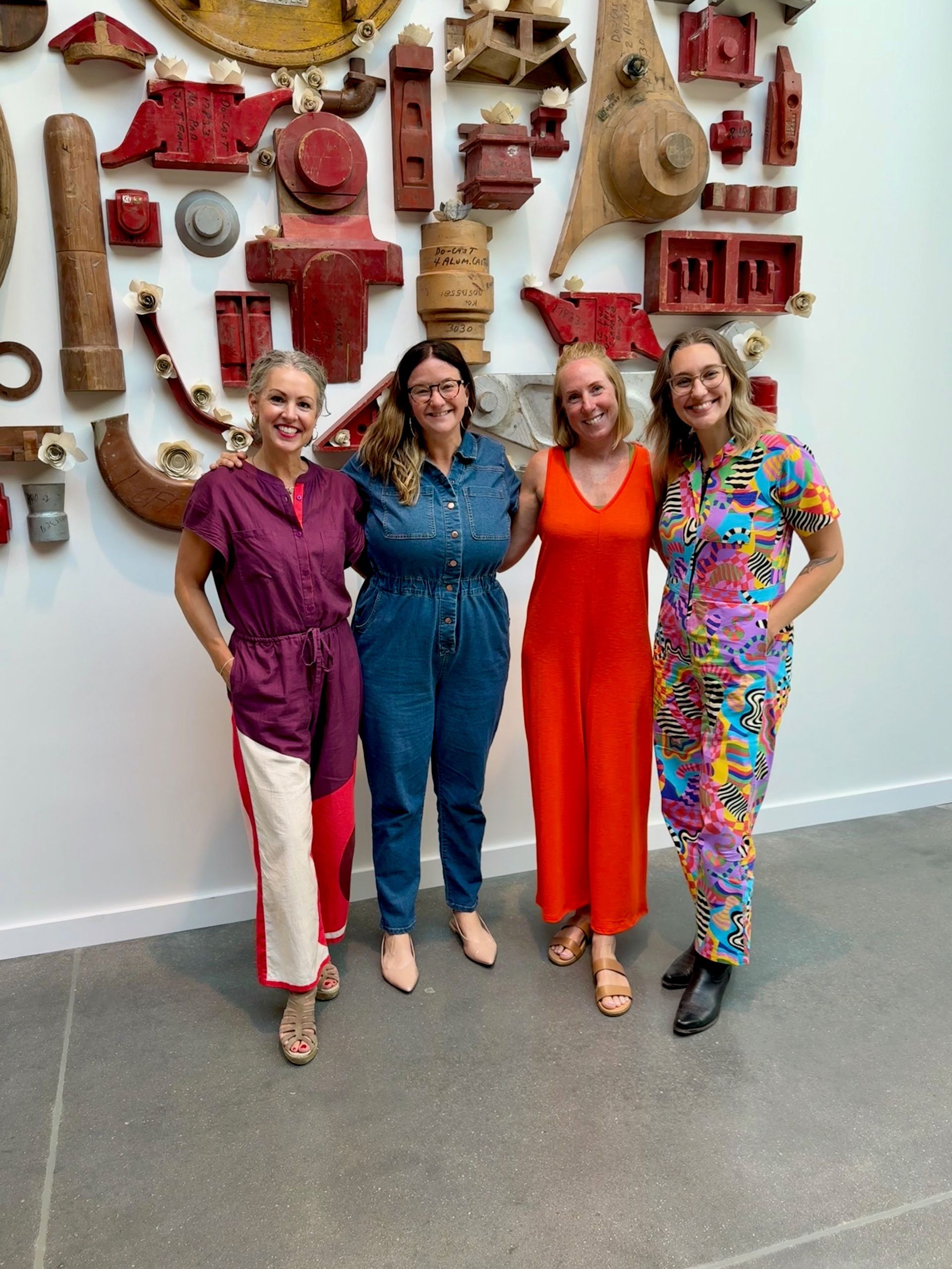 Four women smiling in front of a wall of repurposed art. Red, orange, blue, and patterned outfits. Gallery setting.