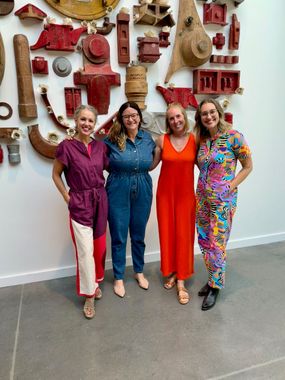 Four women in colorful outfits stand in front of a wall of red and tan industrial art.