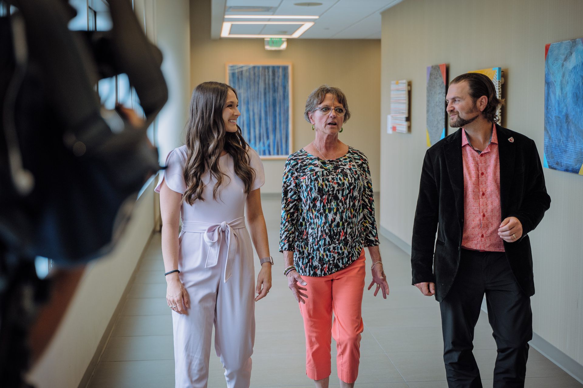 Three people walk down a hallway. A woman in pink pants talks, smiles. A man and woman smile, look on. Artwork hangs.