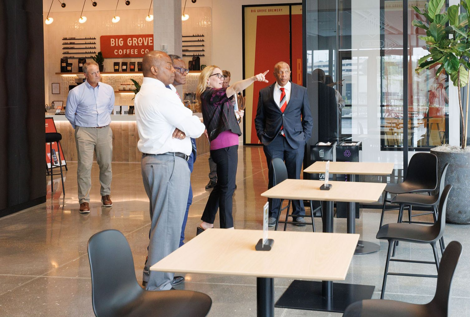 A group of people in a modern cafe, one woman points; others look on near tables.