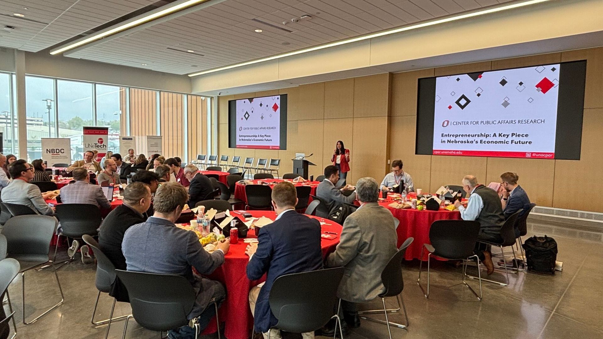 Conference attendees seated at round tables, listening to speakers. Red tablecloths and presentation screens.
