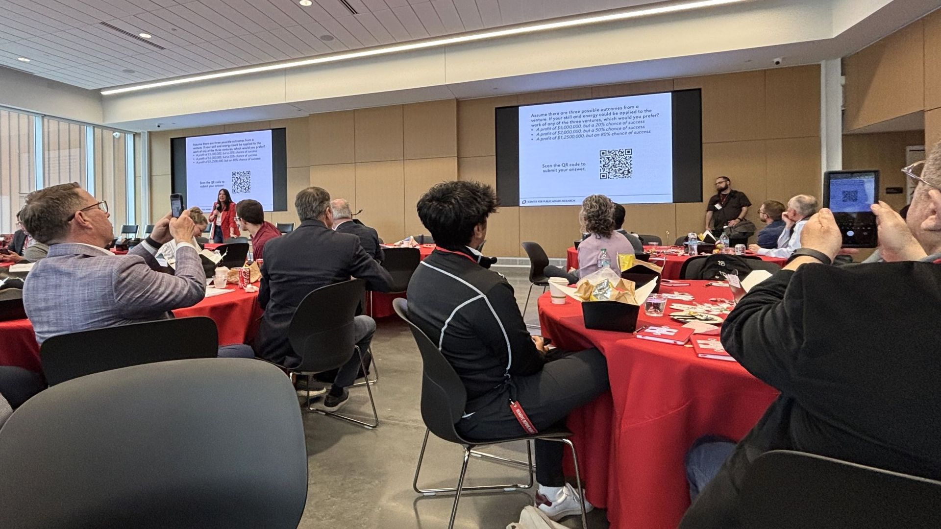 Audience in a conference room with red tablecloths, watching a presentation on screens.