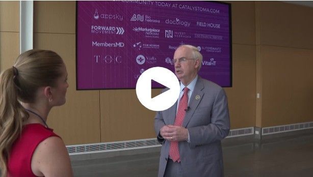 A woman interviews a man in front of a sign with logos. They are in a building.
