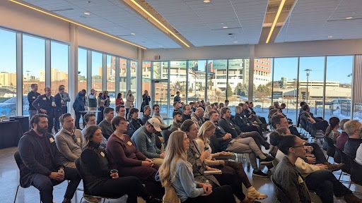 Audience seated, listening in a sunlit room with large windows. People of various ethnicities and ages.