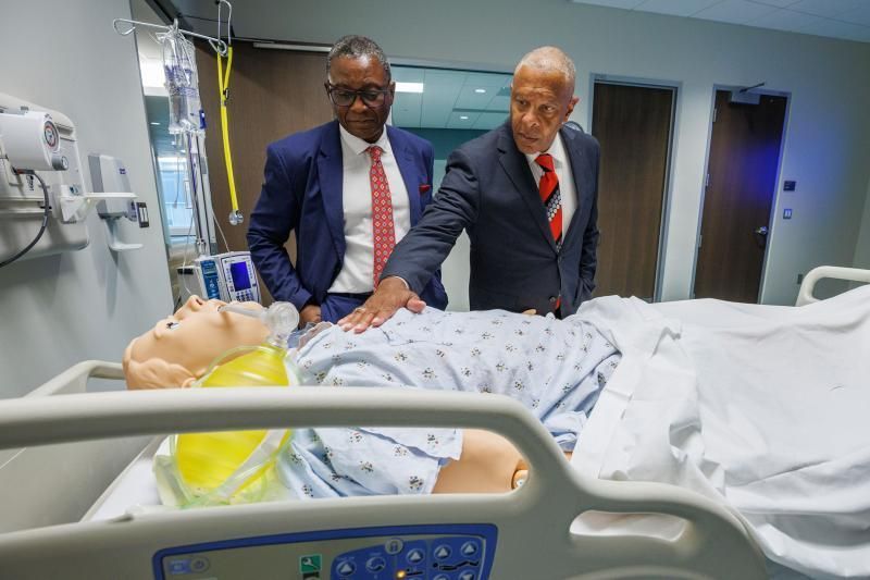 Two men in suits examining a medical simulation mannequin in a hospital room.