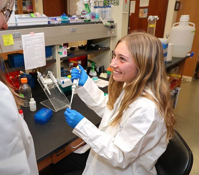 Woman in lab coat uses a pipette, smiling, in a lab setting.