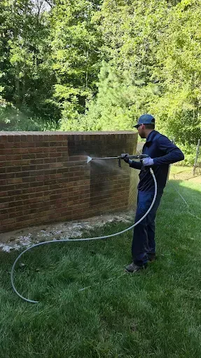Man power washing a brick wall outside. Green grass and trees in background.