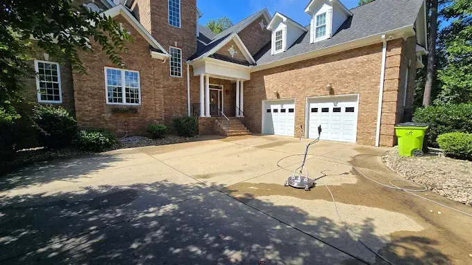 Driveway being pressure washed in front of a two-story brick house with white trim and garage doors.