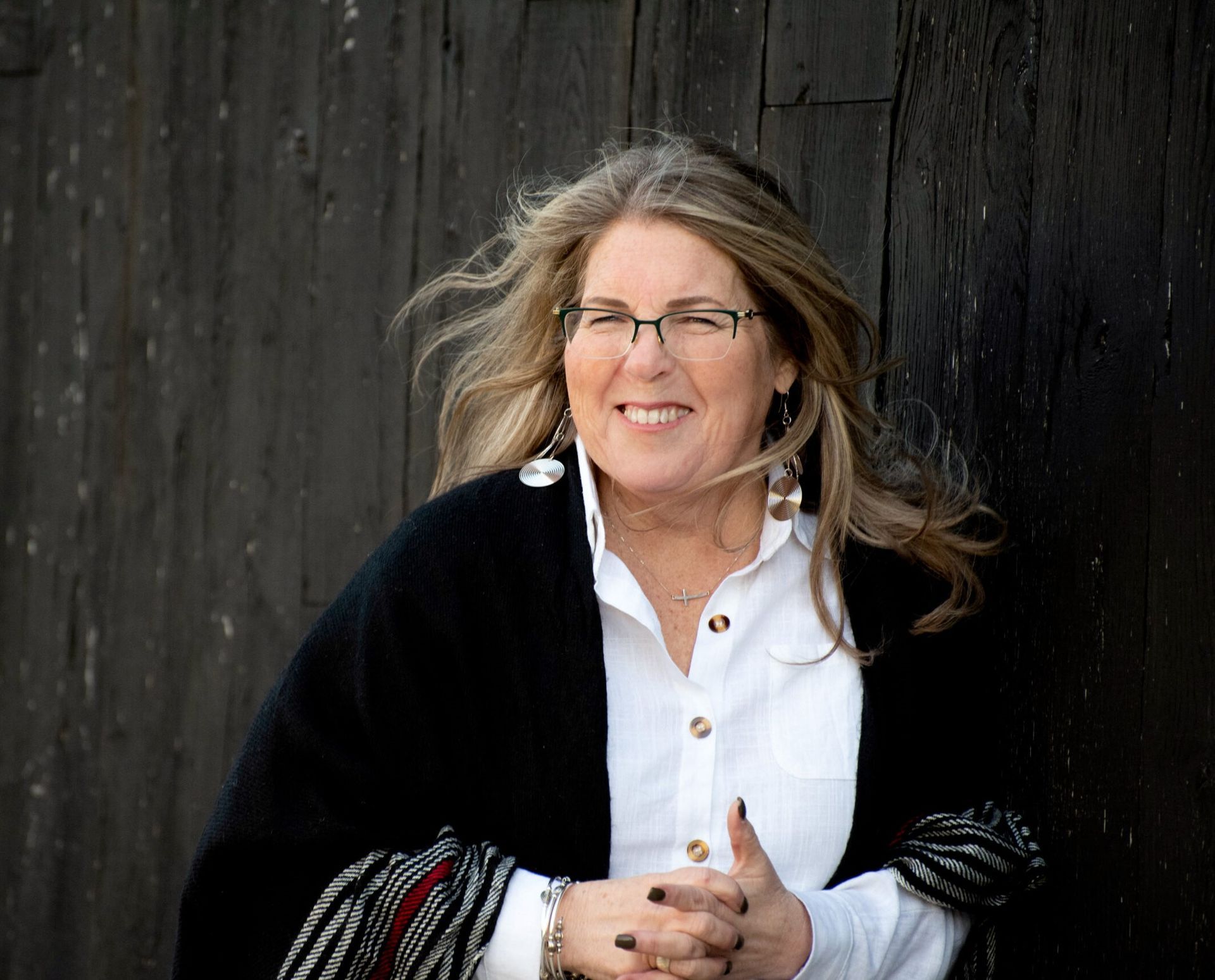 A woman wearing glasses and a white shirt is standing in front of a wooden wall.