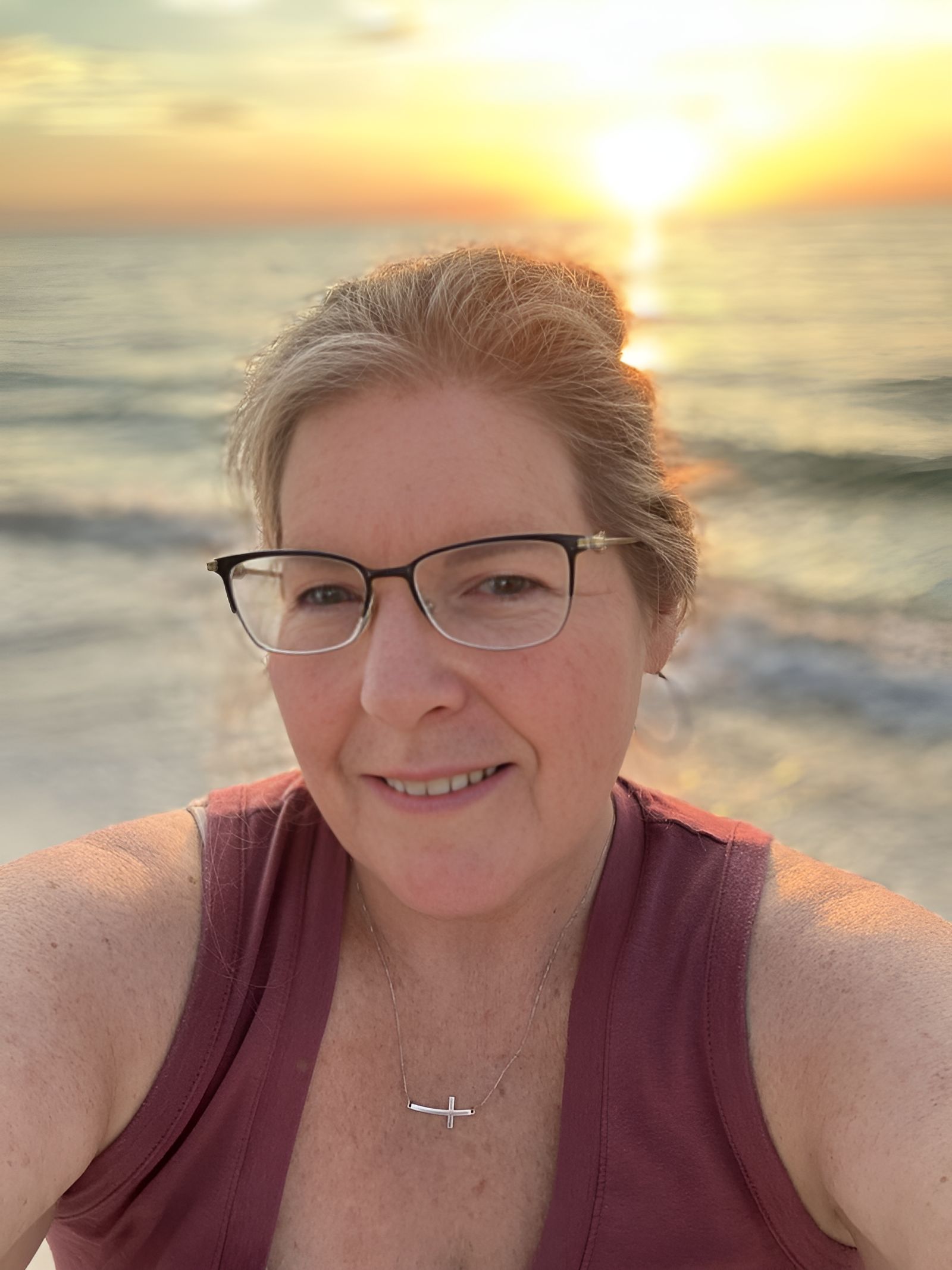 A woman wearing glasses is taking a selfie on the beach at sunset.