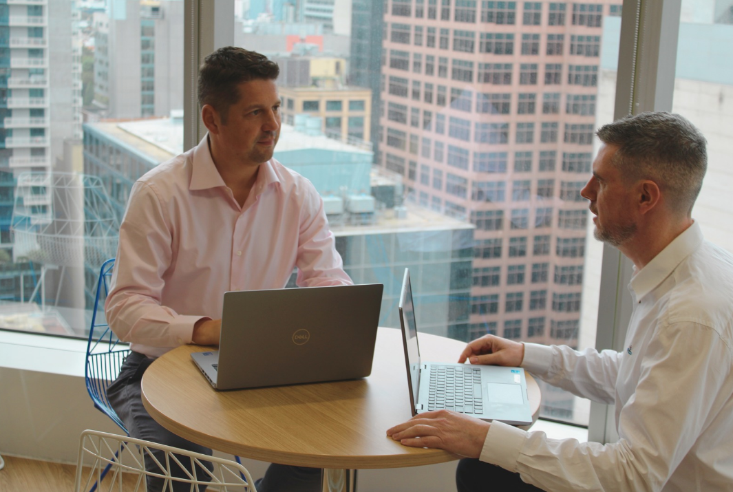 Two men are sitting at a table with laptops and talking to each other.