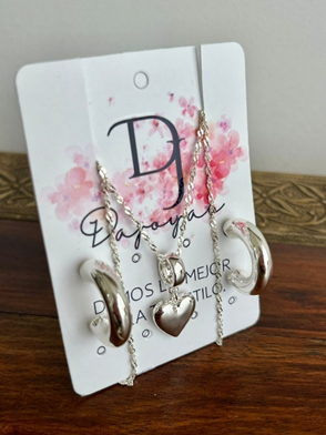 Conjunto de joyería de plata con colgante de corazón y pendientes de aro sobre cartulina blanca con diseño floral rosa.