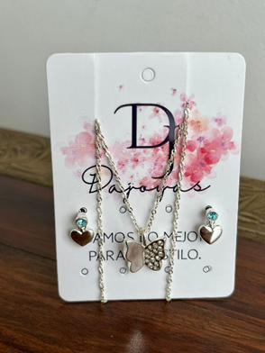 Conjunto de collar y pendientes sobre cartulina blanca, con colgantes de corazón y fondo floral rosa.