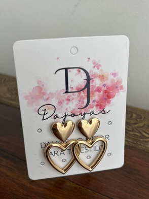 Pendientes dorados en forma de corazón exhibidos sobre una tarjeta blanca con un logotipo, colocada sobre una superficie de madera.